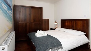 Un dormitorio con cabecera de madera, cama con sábanas blancas, una toalla enrollada sobre la cama, mesita de noche con lámpara y un armario grande.