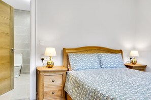 Un dormitorio con una cama de madera, dos mesitas de noche con lámparas, una mesita auxiliar de madera y una pared blanca.