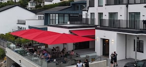 Un espacio al aire libre con mesas, sombrillas rojas, barandillas de vidrio y personas cenando.
