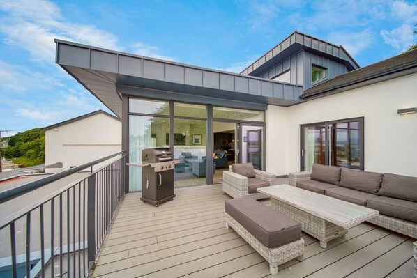 An outdoor deck with a grill, wicker furniture, and a glass door leading to a room. - Bayview Downings - Luxury 4 Bedroom Penthouse (Downings)