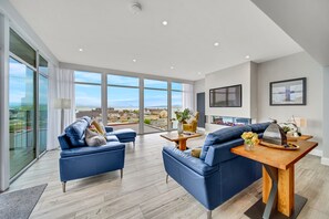 A spacious living area with two blue sofas, a wooden coffee table, and a large window offering a city view. - Bayview Downings - Luxury 4 Bedroom Penthouse (Downings)