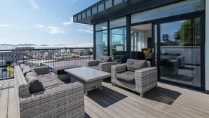 En moderne udendørs terrasse med fletningsmøbler, et glasindrammet område og havudsigt.