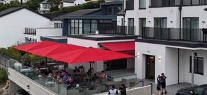 Un espace extérieur aménagé avec des parasols rouges, des garde-corps en verre et des personnes qui prennent leur repas.