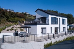 Une maison moderne à deux étages, dotée d’un balcon, avec une voiture garée dans l’allée et un ciel dégagé.