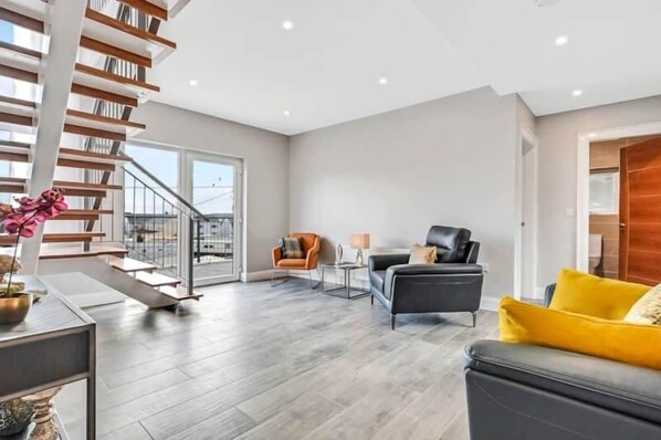 Amplia sala de estar con una escalera moderna, un sofá de cuero negro y un cojín amarillo decorativo.