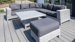 Ein moderner Außenbereich mit einer Eckcouch und Hocker auf einer Holzterrasse, mit Blick auf einen Hügel mit Bäumen und Gebäuden.