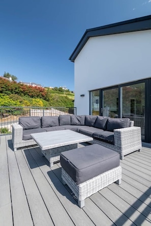 Een modern buitenzitgedeelte met een sectiebank en poufjes op een houten terras, met uitzicht op een heuvel met bomen en gebouwen.