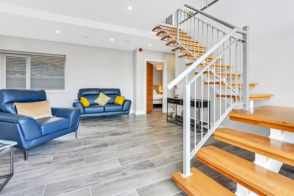 A modern living room with a blue leather sofa, wooden staircase, and grey tiled flooring.