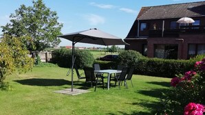A well-maintained backyard with a dining table and chairs under a large umbrella, surrounded by lush greenery and a house in the background. - Quietly located vacation apartment near the dike (Wurster Nordseeküste)