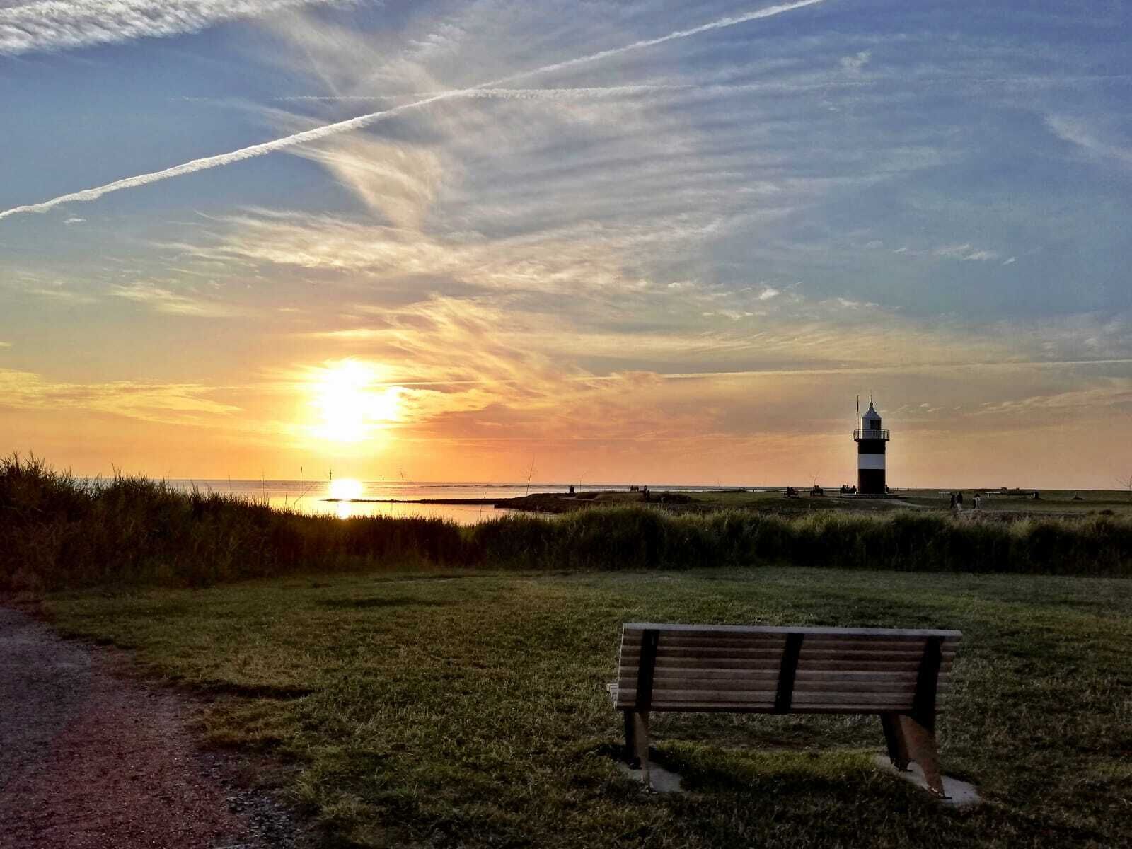 Eine Bank in einem grasbewachsen Feld, im Hintergrund ein Leuchtturm bei Sonnenuntergang.