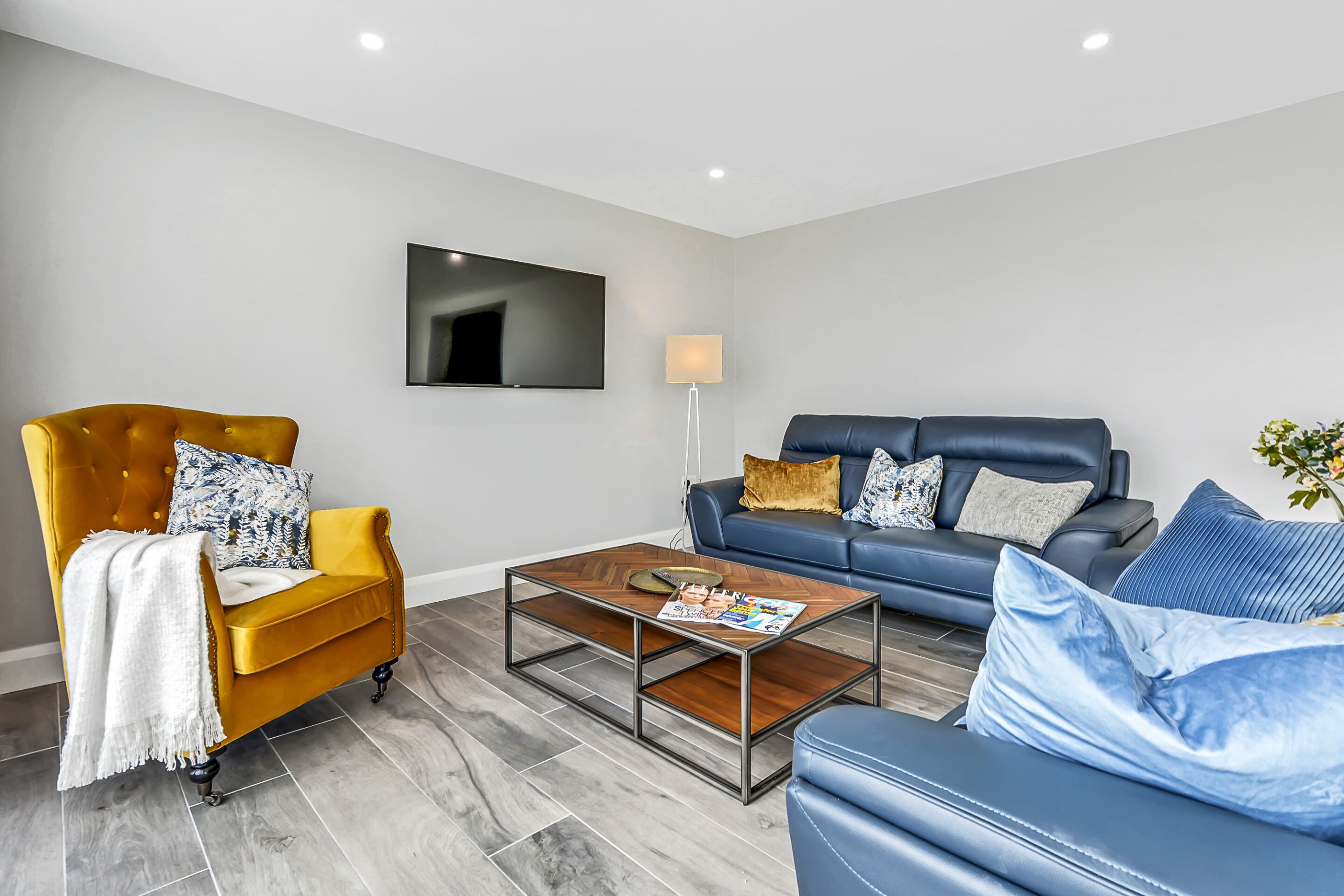 A modern living room with a yellow armchair, a blue sofa, a coffee table, and a flat-screen TV mounted on the wall.