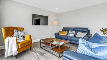 A modern living room with a yellow armchair, a blue sofa, a coffee table, and a flat-screen TV mounted on the wall.