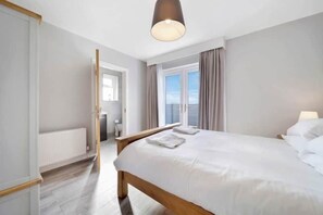 A modern bedroom with a large bed, a wooden headboard, a ceiling light, and a view of the outdoors through a window. - The View, Downings - Luxury House (Downings)