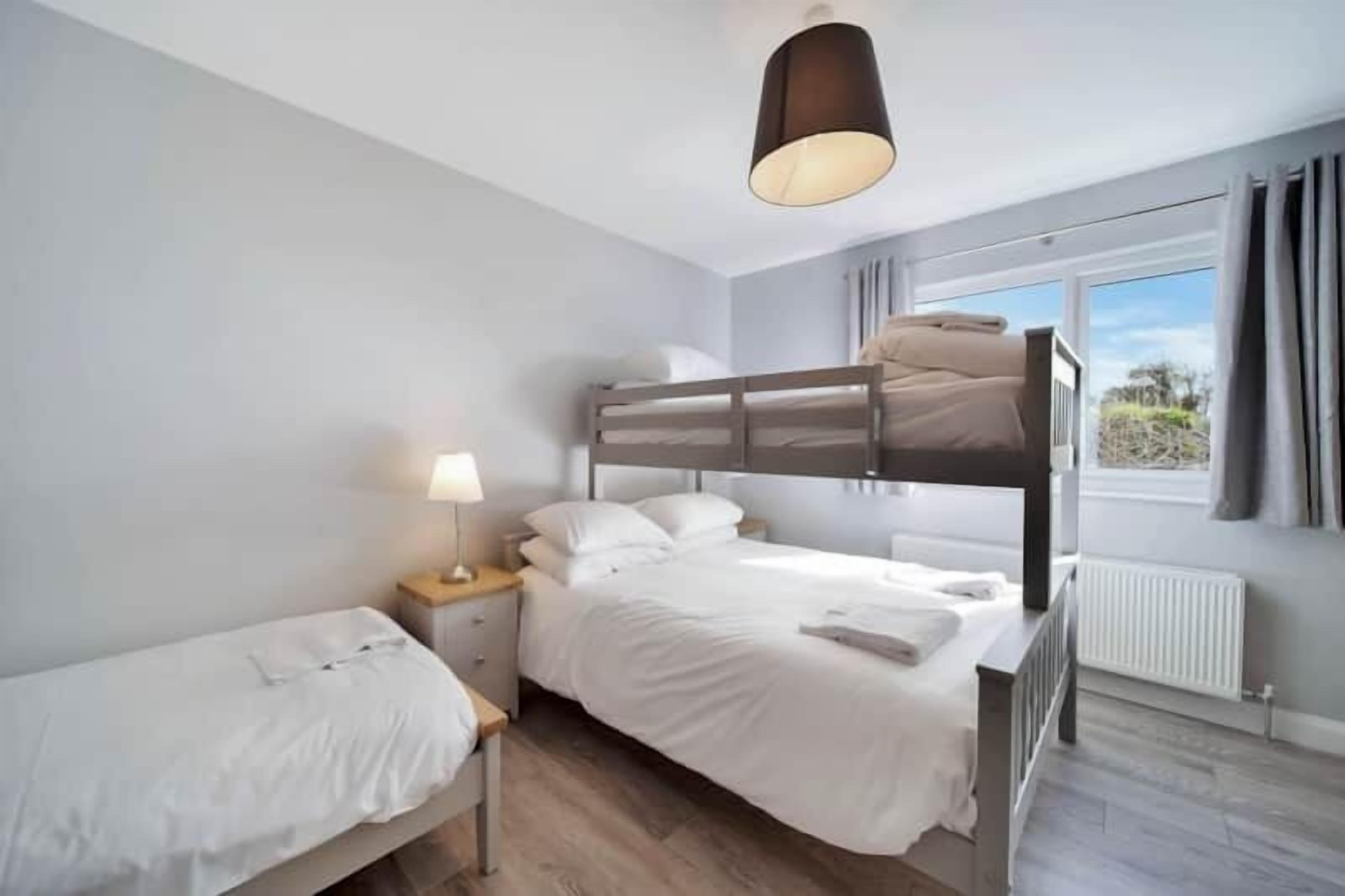 A bedroom with a bunk bed, a single bed, a bedside table with a lamp, and a window with curtains.