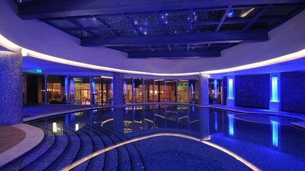 Un espace piscine intérieur moderne, doté d’un design aux lignes courbes, de murs en verre et d’un éclairage d’ambiance bleuté.