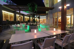 Un espacio de comedor al aire libre con mesas y sillas, un área de estar cubierta y un edificio con ventanas y puerta amplias.