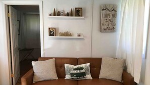 Ein Wohnzimmer mit einem braunen Ledersofa, zwei gemusterten Kissen, einem gerahmten Bild und zwei Regalen mit Dekorationsobjekten und einem Schild.