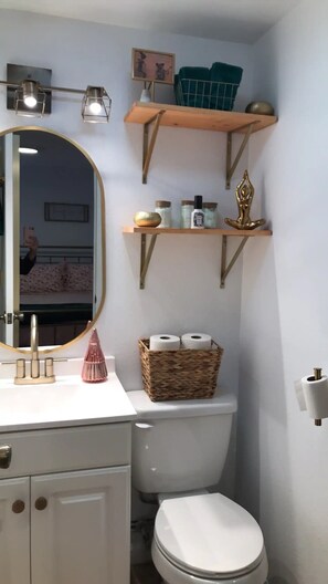 Ein Badezimmer mit einer weißen Toilette, einem runden Spiegel mit goldener Rahmung, einem Waschtisch mit weißer Arbeitsplatte und zwei oberhalb der Toilette angebrachten Ablagen mit Pflegeprodukten und einem dekorativen Gegenstand.