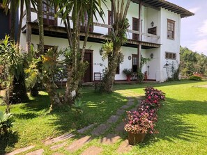Um edifício de dois andares com fachada branca e detalhes marrons, cercado por vegetação exuberante e um gramado bem cuidado.