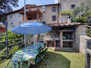 A patio with a table and chairs, a blue umbrella, and a stone building with a chimney in the background.