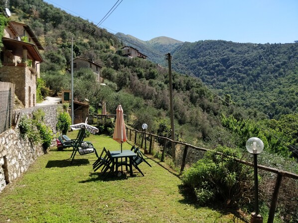 A hillside property with a stone wall, a grassy area with outdoor furniture, and a view of the surrounding forested hills.