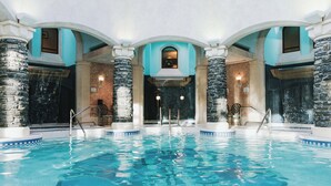 An indoor swimming pool with stone pillars, a view of the sky through an arched opening, and a waterfall feature. - Fairmont Banff Springs Gold Experience (Banff)