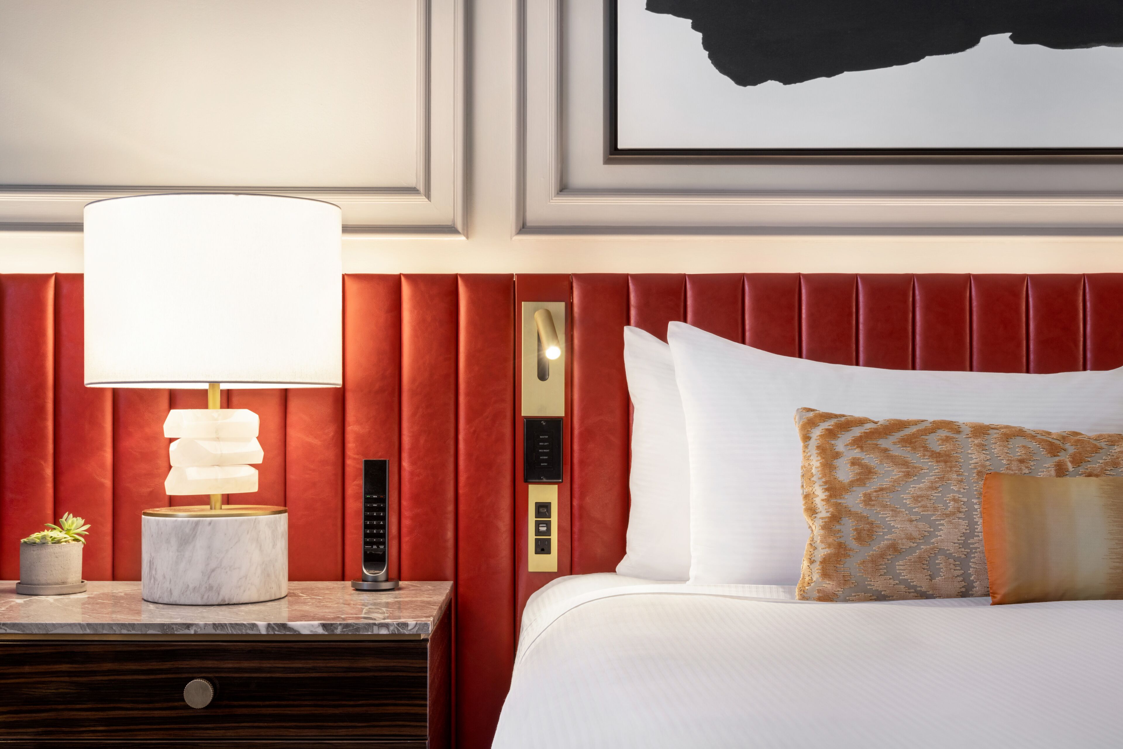 Habitación de hotel con cabecera tapizada en rojo, mesita de noche de mármol con lámpara, planta en maceta pequeña y cama con ropa de cama blanca y almohadas estampadas.