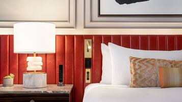 Habitación de hotel con cabecera tapizada en rojo, mesita de noche de mármol con lámpara, planta en maceta pequeña y cama con ropa de cama blanca y almohadas estampadas.