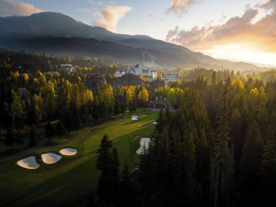 A golf course with bunkers, surrounded by dense forest and mountains in the background.
