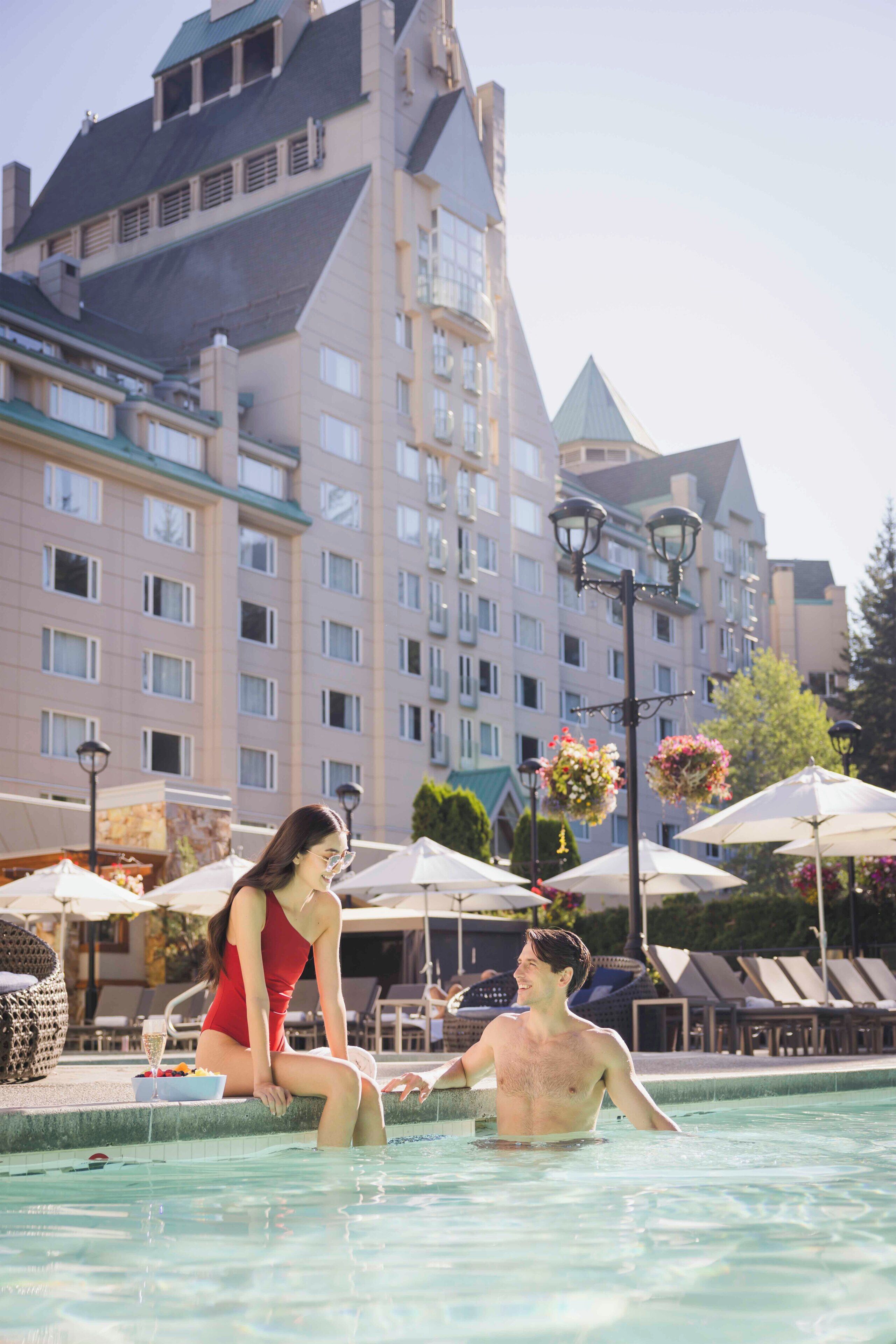 A swimming pool with lounge chairs and umbrellas, a building with a distinctive tower, and a person sitting by the pool.