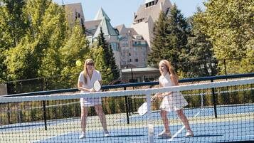 A tennis court with a net, two players, and a building with a green roof in the background.