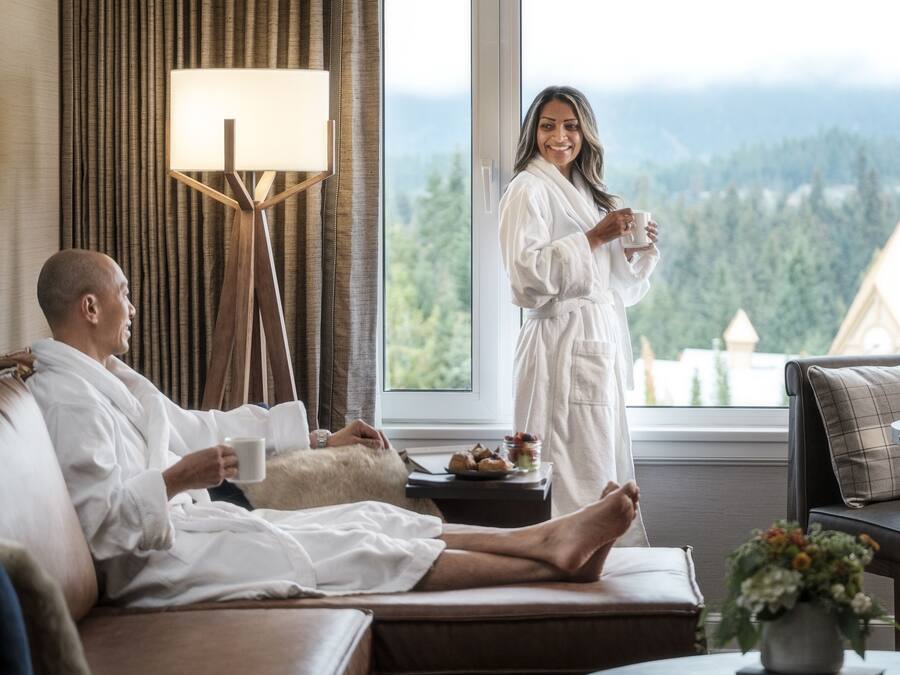 A hotel room with a large window offering a view of a forested mountain range. There is a bed with two people in white bathrobes, a standing person holding a cup, and a small table with a plant and a tray of food.