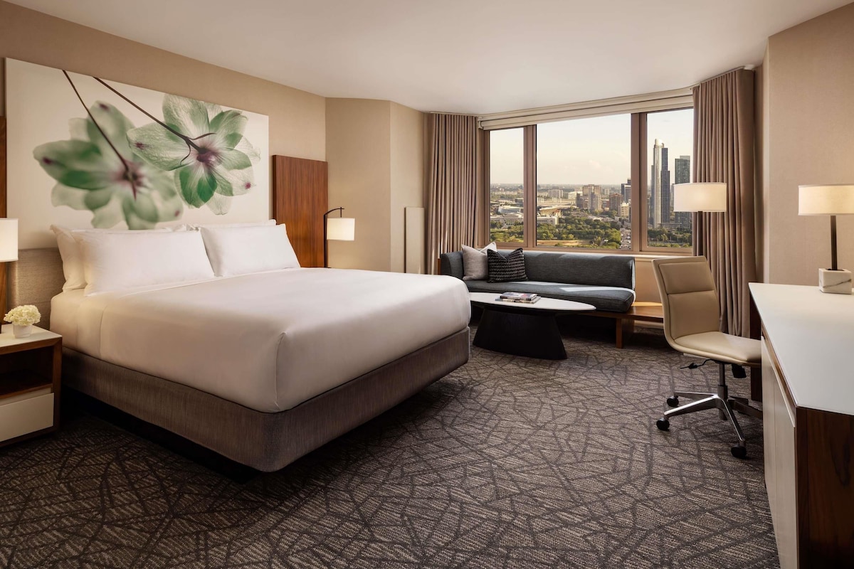 Une chambre d’hôtel avec un lit, un canapé, un bureau et une grande fenêtre donnant sur la ville.