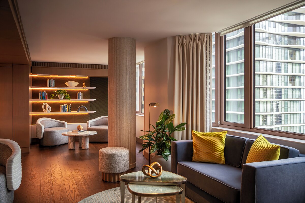 Un salon moderne comprenant un canapé, des fauteuils, une table basse, une bibliothèque et une grande fenêtre donnant sur des immeubles.