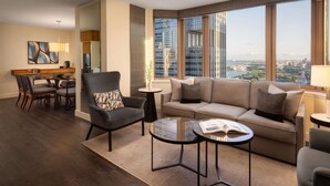 A modern hotel room with a dining area, a sofa set, a small table with a book, and large windows offering a city view. - Fairmont Chicago at Millennium Park Gold Experience (Chicago)