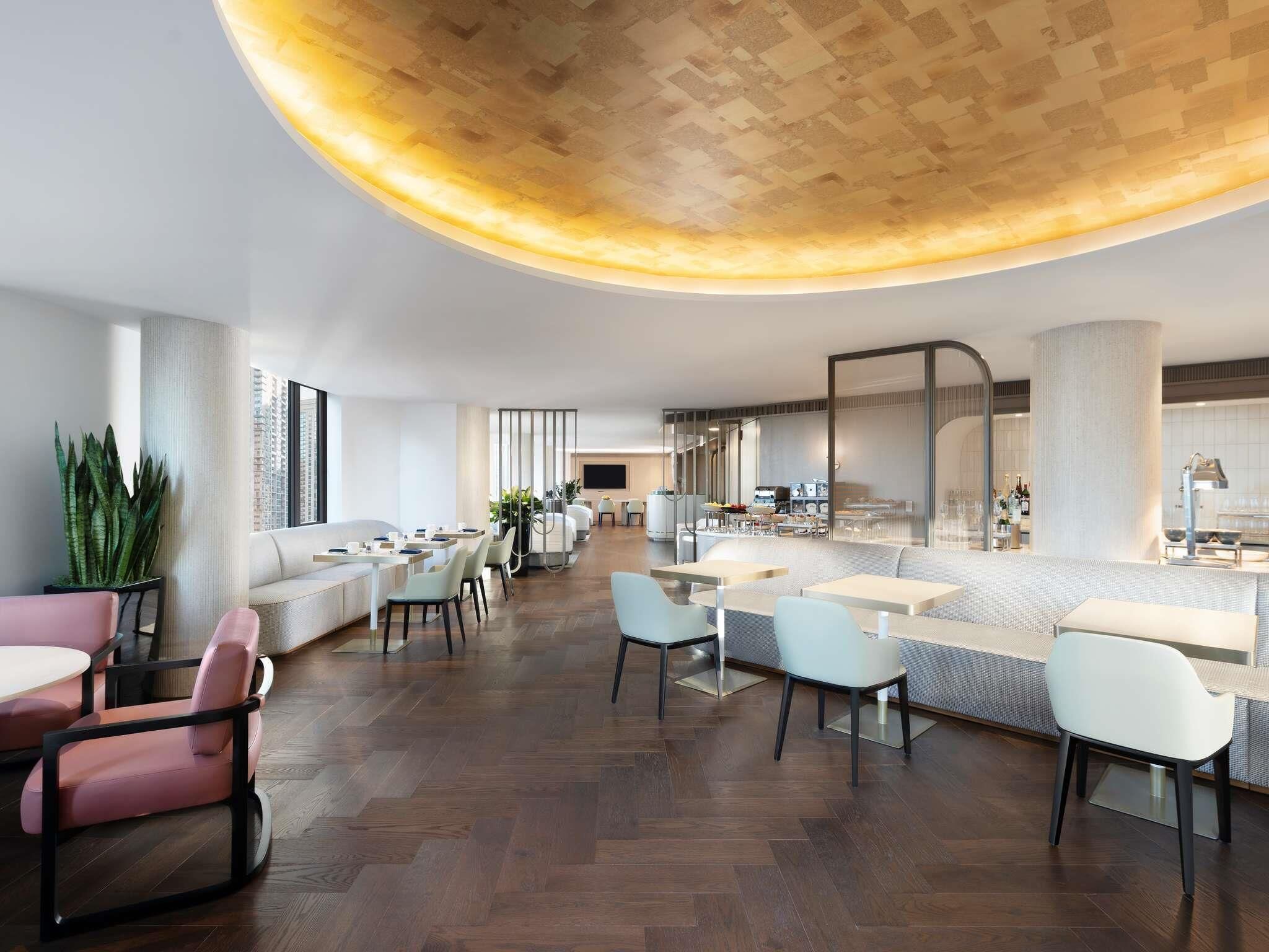 Una hall moderna di un hotel con soffitto curvo, zone relax, un bar e una cucina visibile sullo sfondo.
