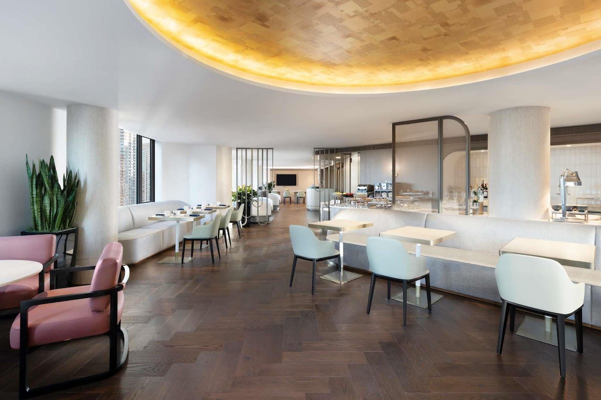 A modern hotel lobby with a curved ceiling, seating areas, a bar, and a kitchen visible in the background.