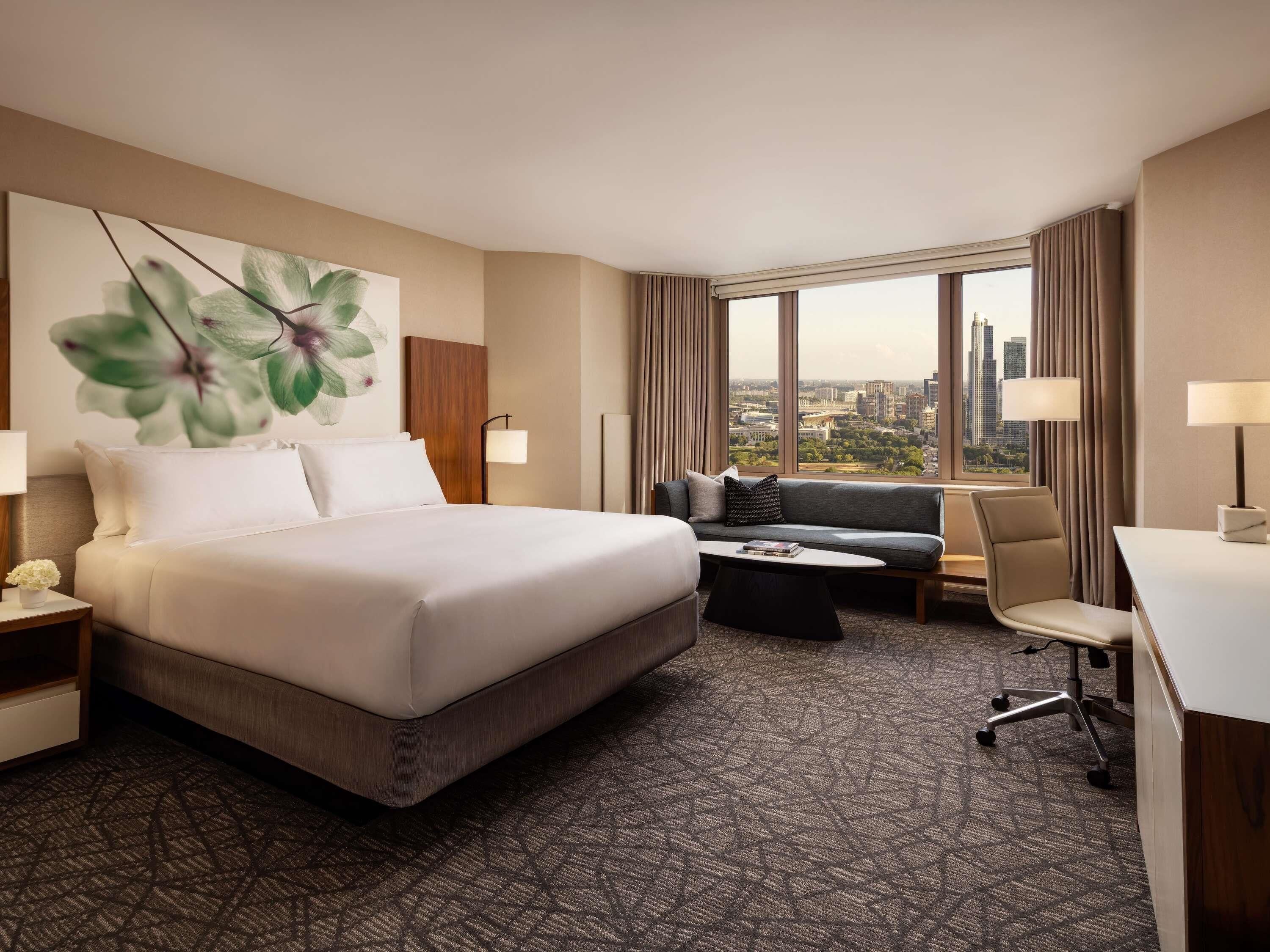 Een hotelkamer met een bed, een slaapbank, een bureau en een groot raam met uitzicht op de stad.