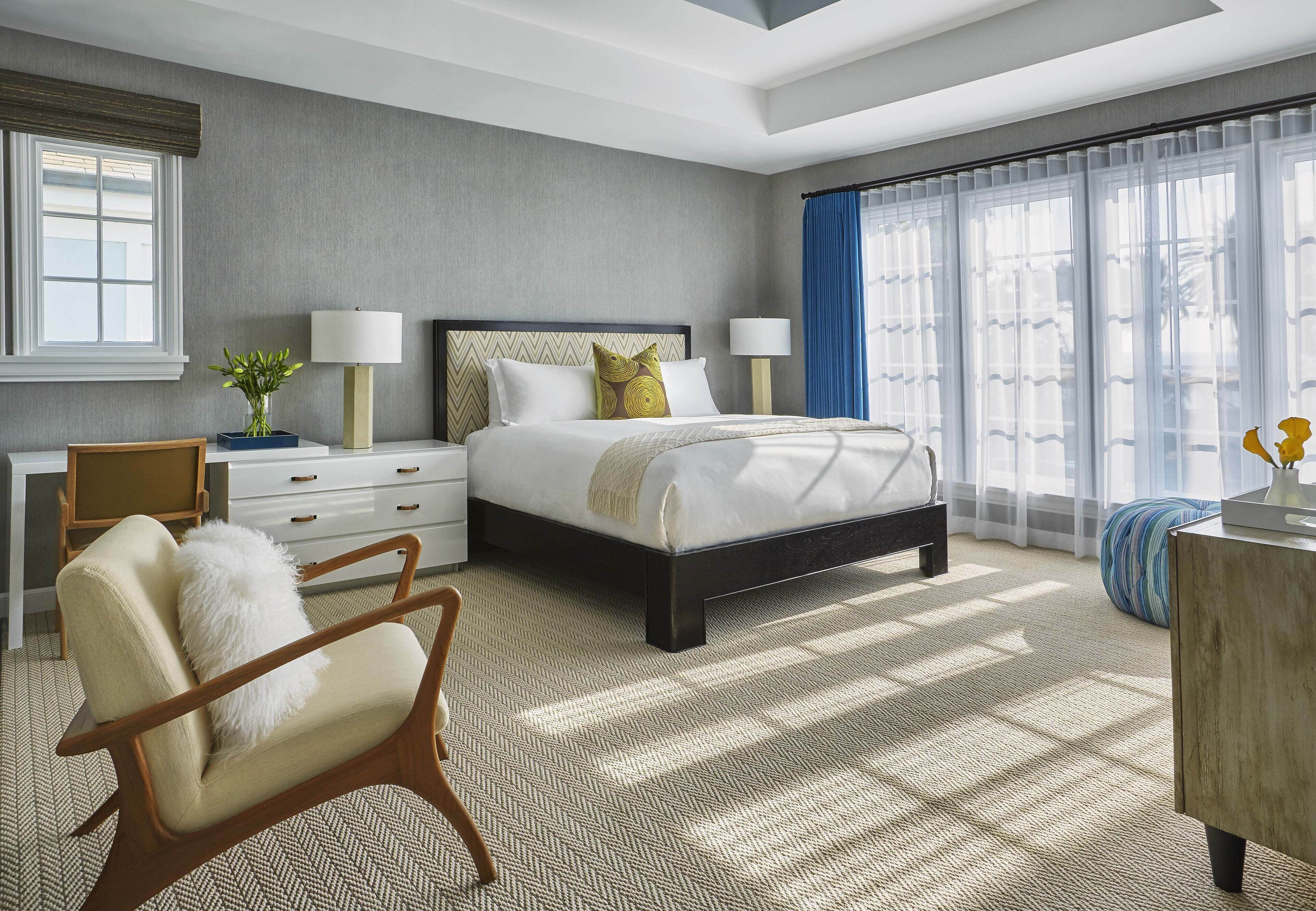 Une chambre d’hôtel moderne dotée d’un grand lit, de deux tables de chevet, d’une chaise, d’un bureau et d’une grande fenêtre avec des rideaux.