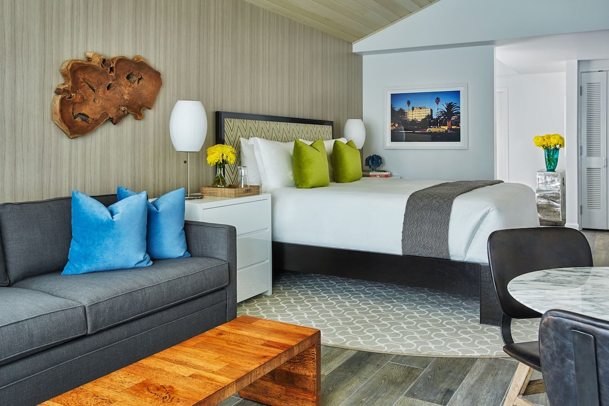 A modern hotel room with a grey sofa, a wooden coffee table, a bed with white and green bedding, a nightstand with a lamp, and a framed picture on the wall.