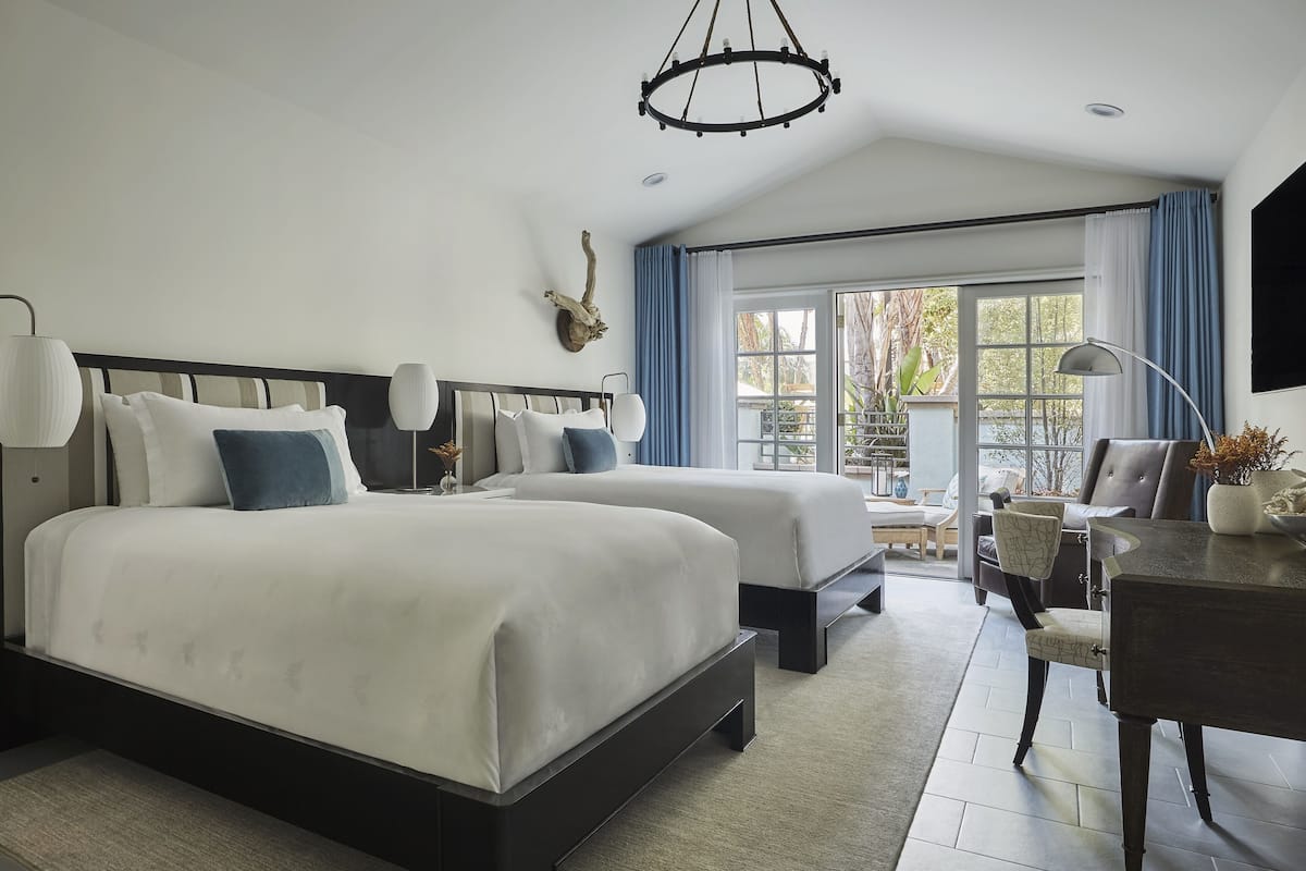 A modern hotel room with two beds, a desk, and a large window offering a view of the outdoors.