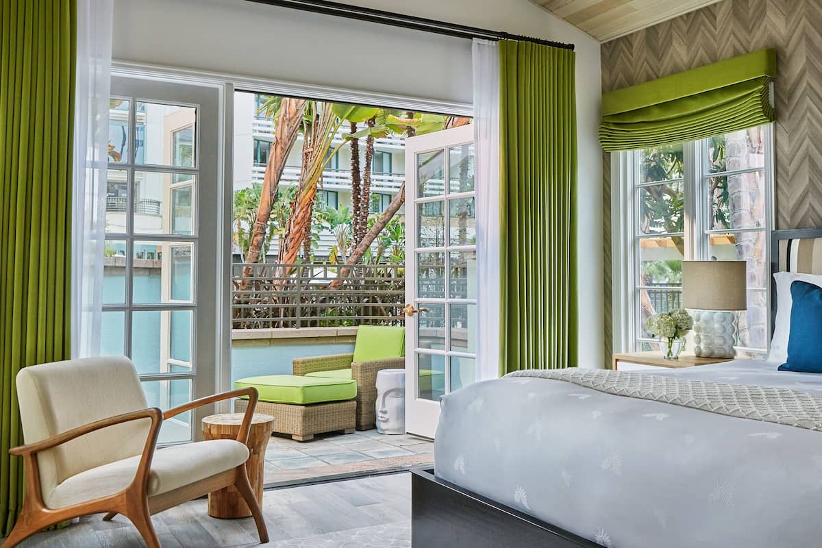 A hotel room with a large bed, a chair, a small table, and a view of a pool and palm trees.