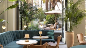 A modern lounge area with a green sofa, round tables, and chairs, a large potted plant, and a view of the outdoor patio. - Fairmont Miramar Bungalows (Santa Monica)