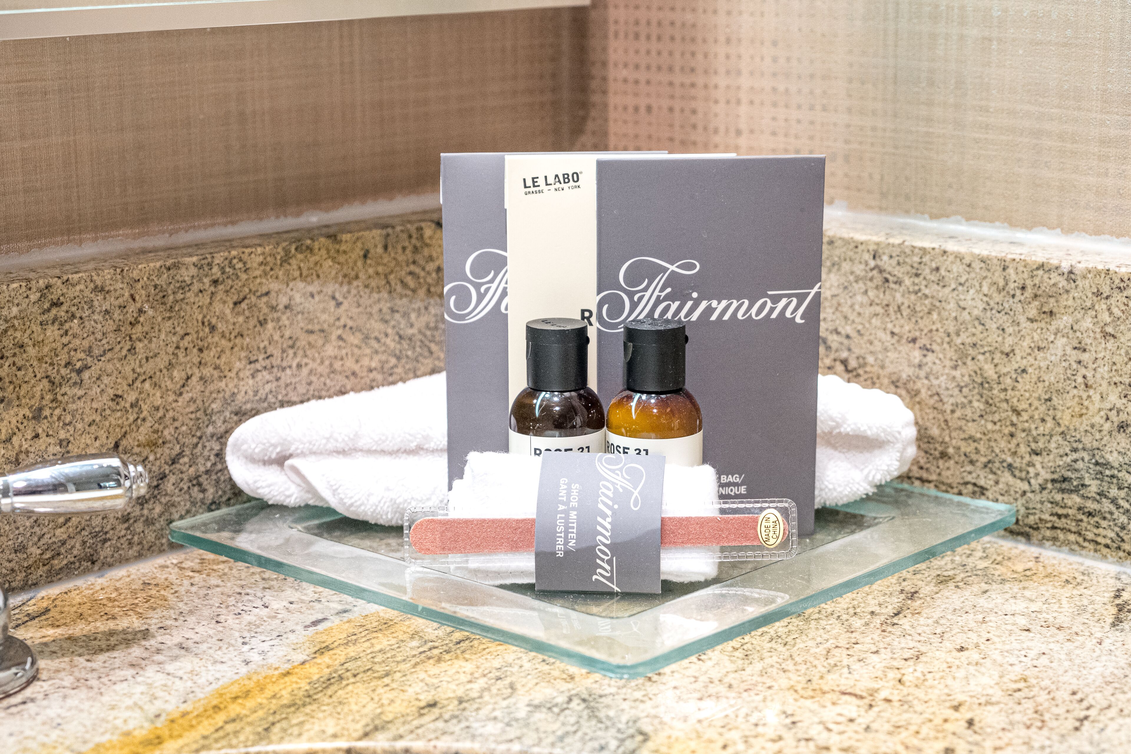 A bathroom counter with a glass tray holding a Le Labo fragrance box, two bottles of perfume, and a white towel.