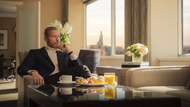 Un hombre sentado a la mesa desayunando, con vista a la ciudad y un jarrón de flores.