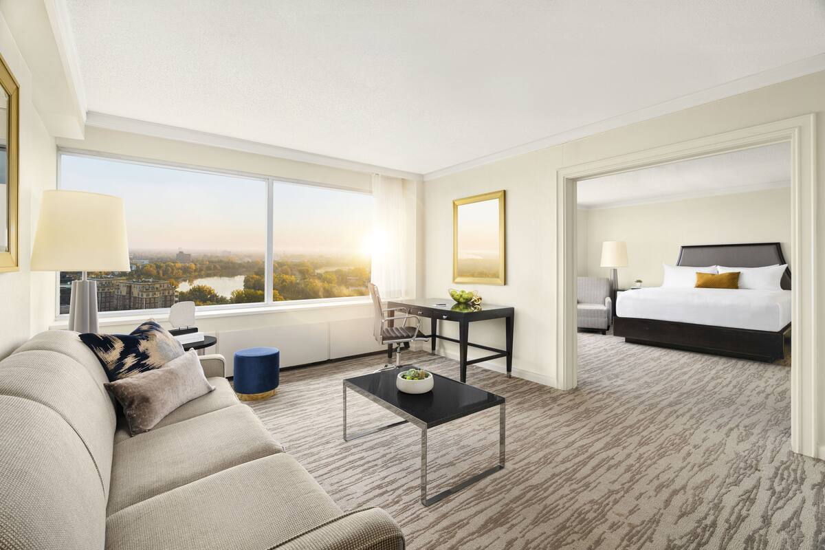 A hotel room with a large window, a sofa, a coffee table, a desk, and a bed.