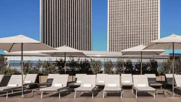 A poolside area with lounge chairs and umbrellas, a clear blue sky, and two modern buildings in the background.