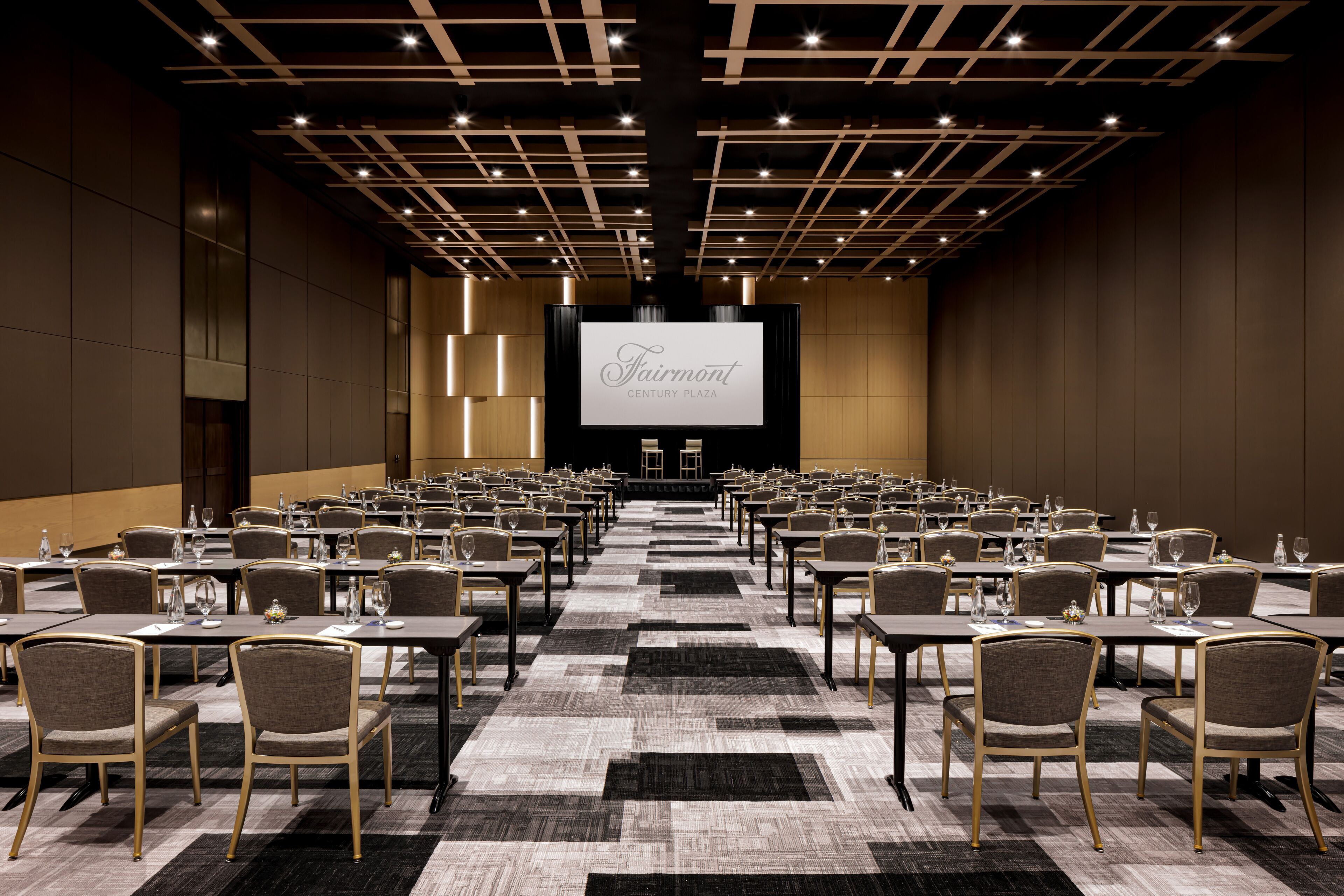 A conference room with rows of tables and chairs, a large screen at the front, and a modern ceiling design.