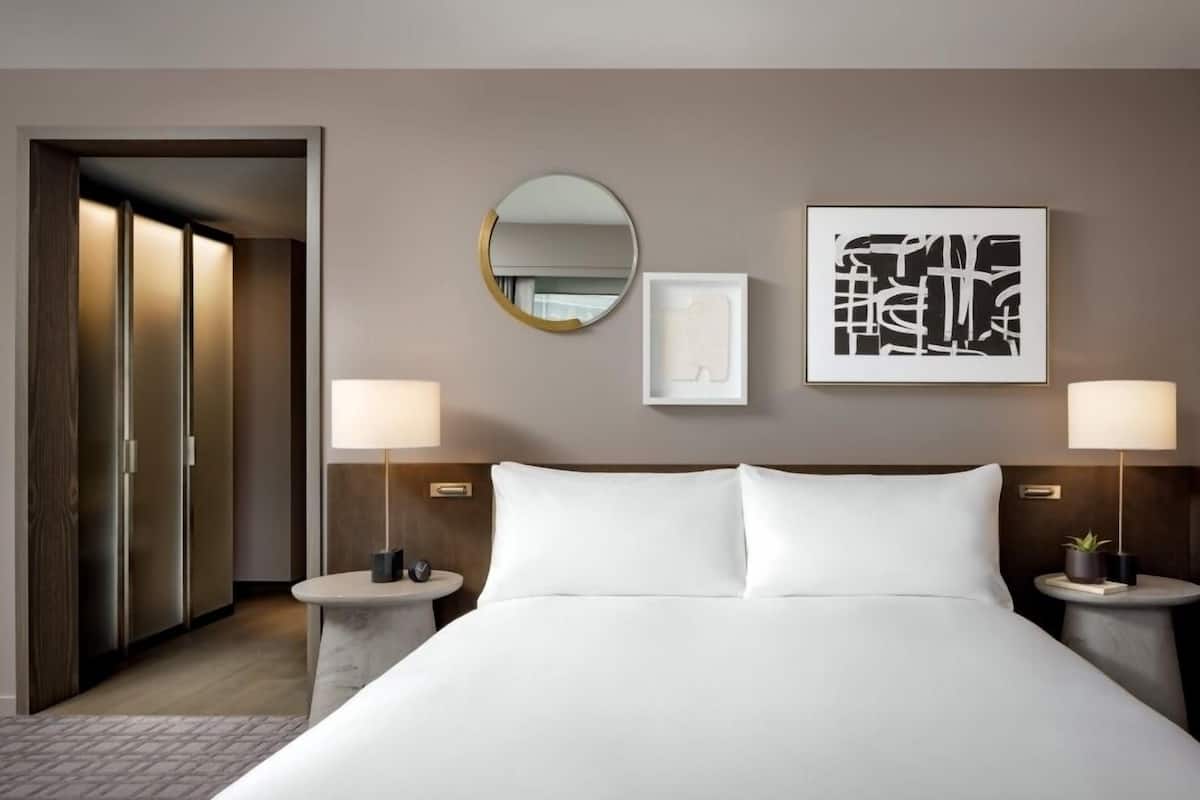 Une chambre d’hôtel avec un grand lit, deux tables de chevet avec des lampes, un miroir rond et des œuvres d’art accrochées au mur.