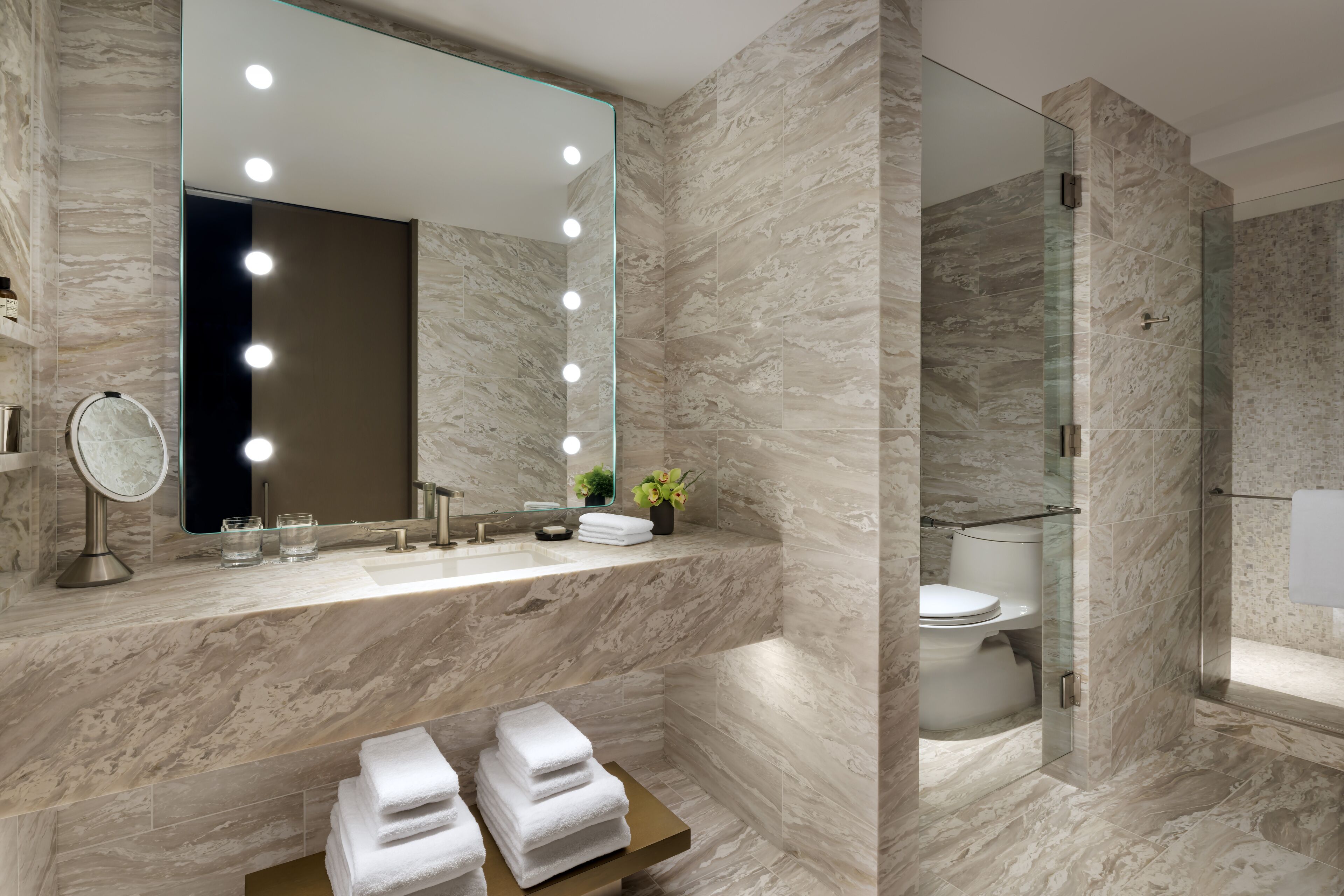 A modern bathroom with a marble vanity, a large mirror with lights, a glass shower enclosure, and neatly stacked towels.